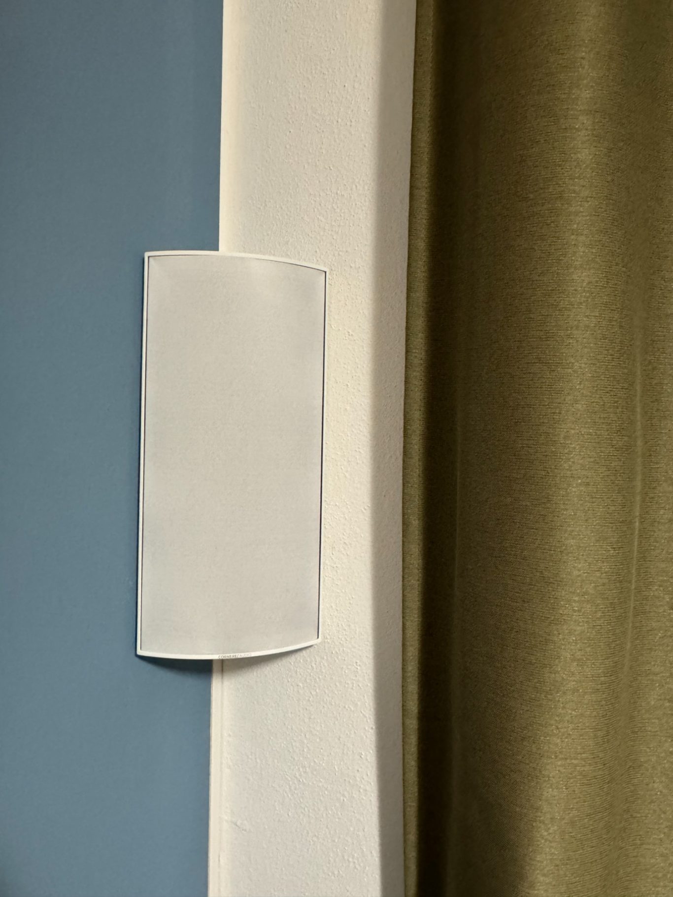 Cornered Audio C3 Weißer Lautsprecher an einer Ecke, neben einem grünen Vorhang und einer petrol Wand.
